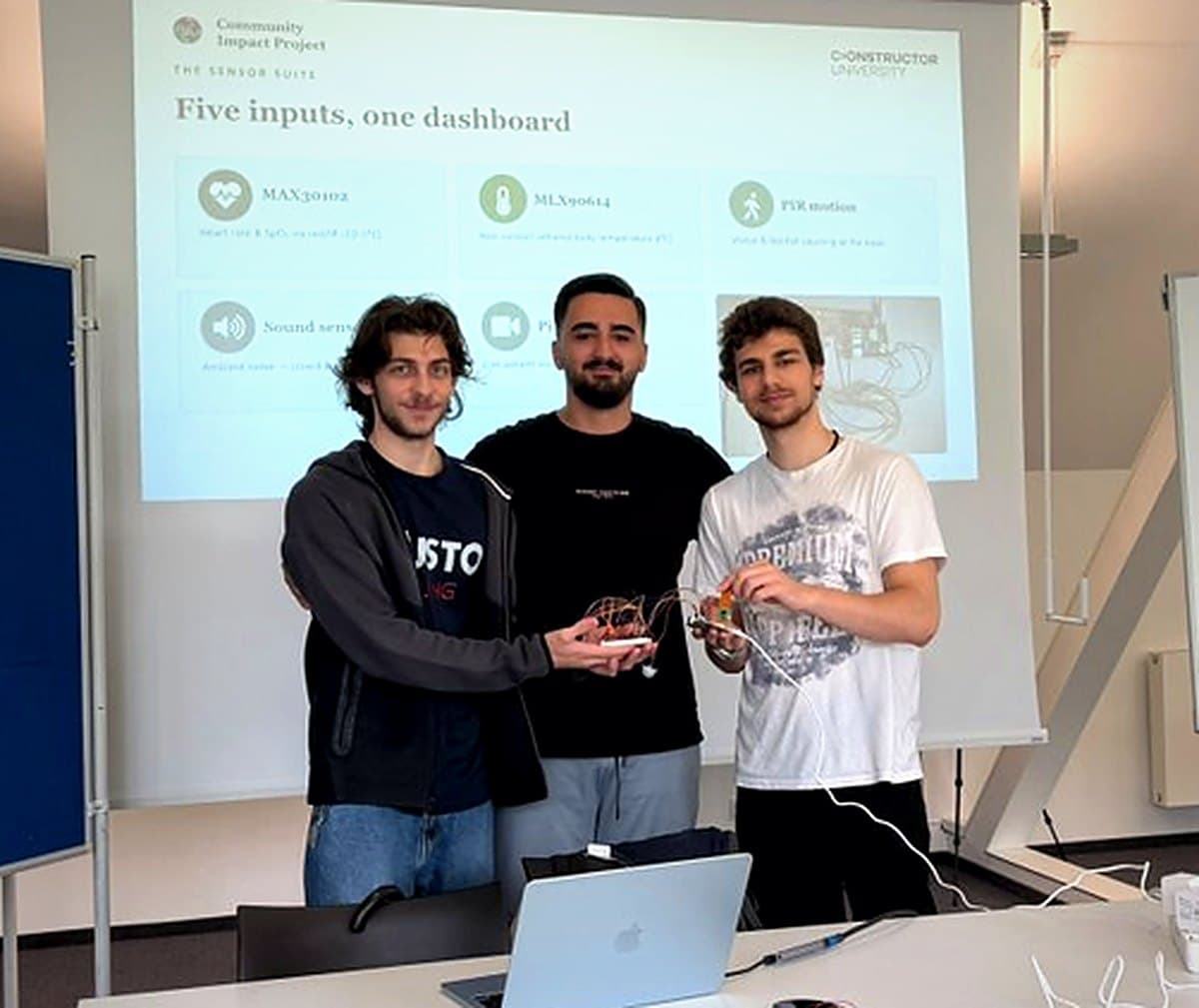دانیال (وسط) و دو شرکتکننده دوره با سنسورهای پروژه Community Impact جلوی اسلاید «Five inputs, one dashboard»